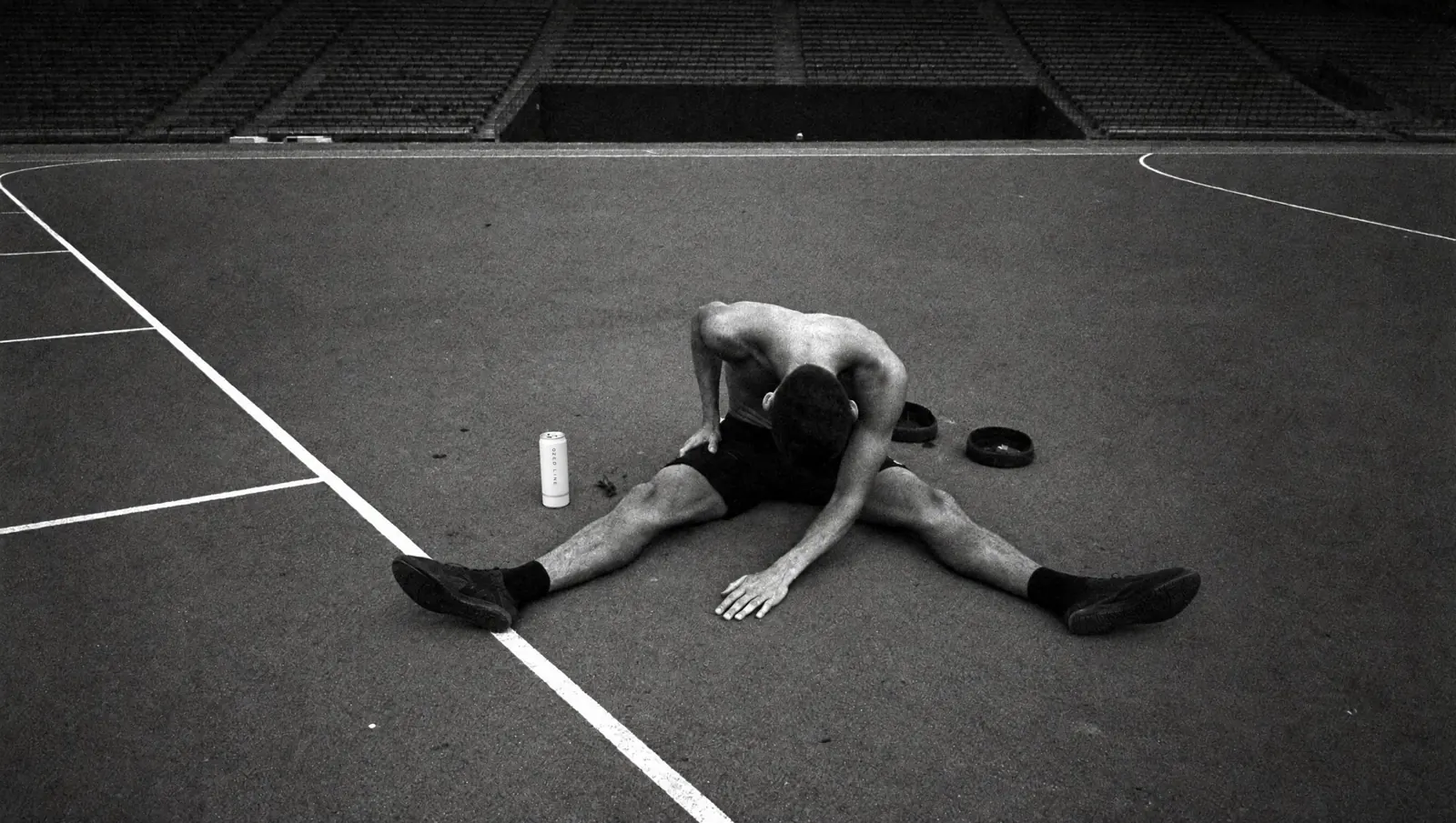 Atleta agotado sentado en una pista de atletismo vacía, con una lata Ozed Line al lado, en blanco y negro